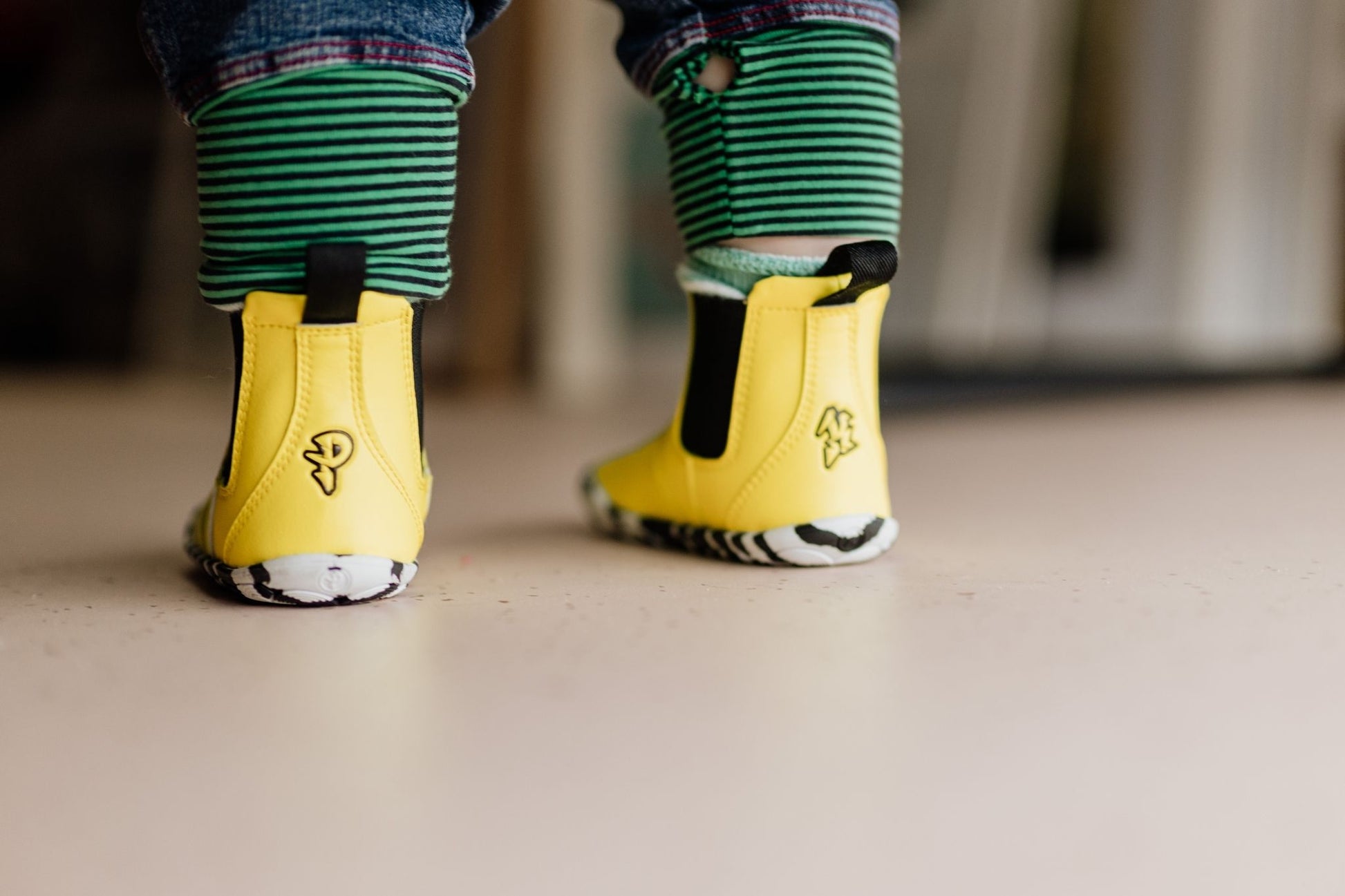 A close-up of a child's feet wearing bright yellow vegan chelsea boots with black elastic side panels and green-and-black striped pant cuffs, standing on a light beige floor.