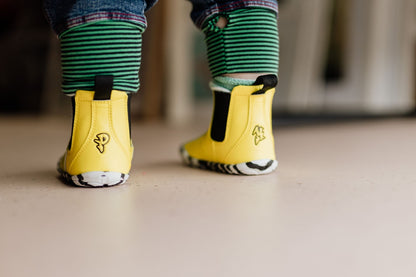 A close-up of a child's feet wearing bright yellow vegan chelsea boots with black elastic side panels and green-and-black striped pant cuffs, standing on a light beige floor.