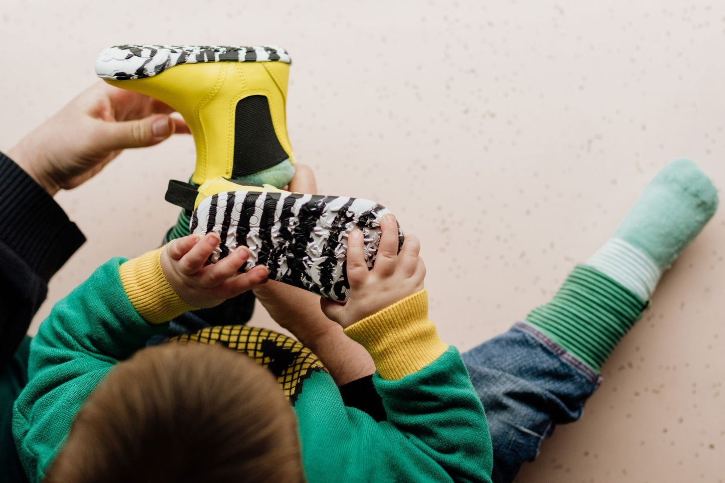 A child wearing a green and yellow sweatshirt and blue jeans lies on a light beige floor while an adult hands them a bright yellow barefoot PaperKrane chelsea boot with a black-and-white zebra print. The child also wears green and mint striped socks.
