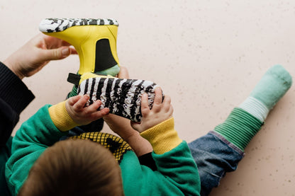 A child wearing a green and yellow sweatshirt and blue jeans lies on a light beige floor while an adult hands them a bright yellow barefoot PaperKrane chelsea boot with a black-and-white zebra print. The child also wears green and mint striped socks.
