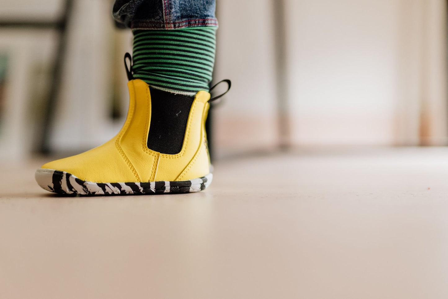 A child's yellow chelsea boot with a black elastic side panel and a black-and-white zebra-patterned sole, worn with green and black striped blue jeans.
