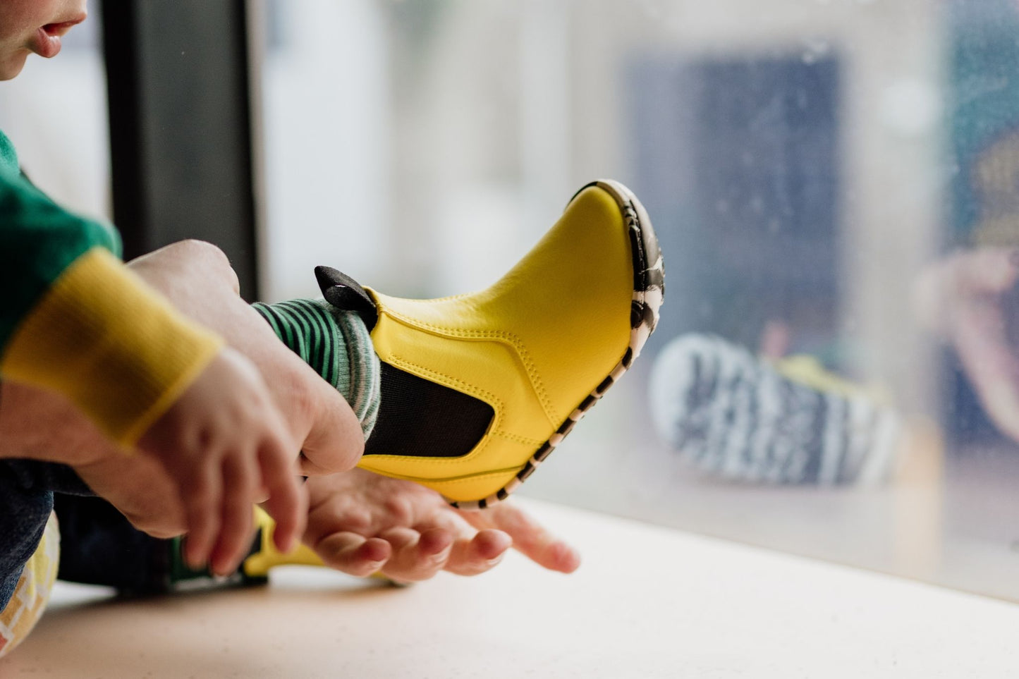 A close-up lifestyle shot of a person sitting by a window, putting on a bright yellow slip-on boot. The toddler is wearing a green top and striped cuffed pants, and the background is a softly blurred window and indoor setting.