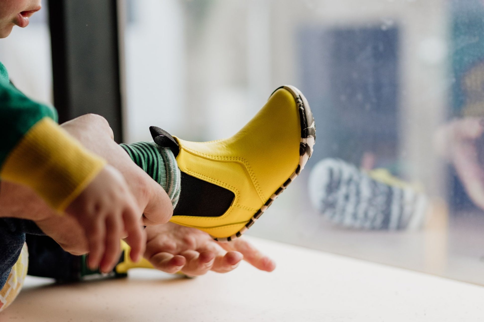 A close-up lifestyle shot of a person sitting by a window, putting on a bright yellow slip-on boot. The toddler is wearing a green top and striped cuffed pants, and the background is a softly blurred window and indoor setting.
