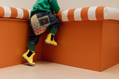 A child wearing a green top, patterned denim pants, with green striped cuffs, and bright yellow PaperKrane barefoot boots attempts to climb on an orange and white striped bench in a minimalist indoor setting.