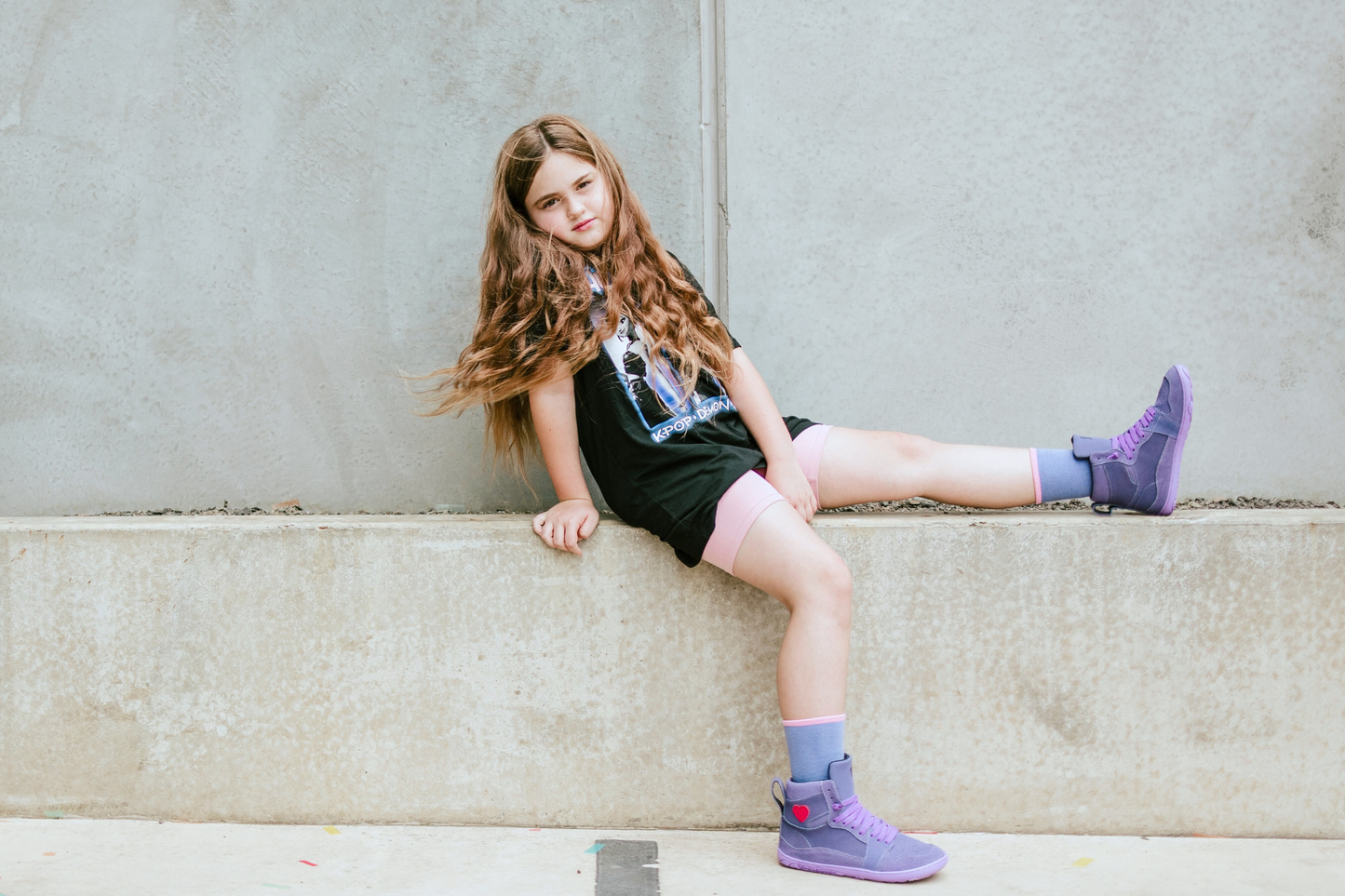 Young girl sitting on a concrete ledge wearing black outfit and purple boots.