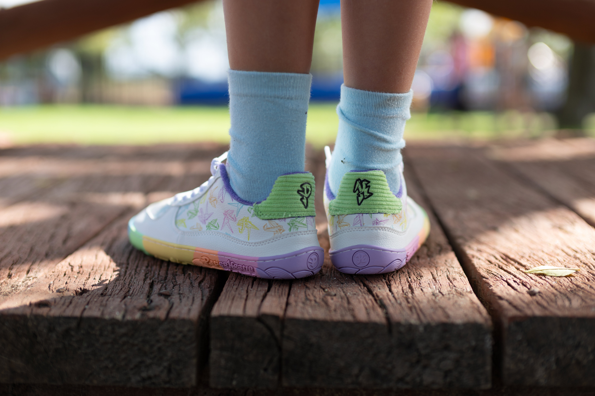 White barefoot sneakers from PaperKrane with a zero drop rainbow sole, and rainbow origami cranes printed on them. 
Seen from the back, there's green corduroy with PK logo details. This pair is being worn by a child with white socks on. On a wooden bridge.