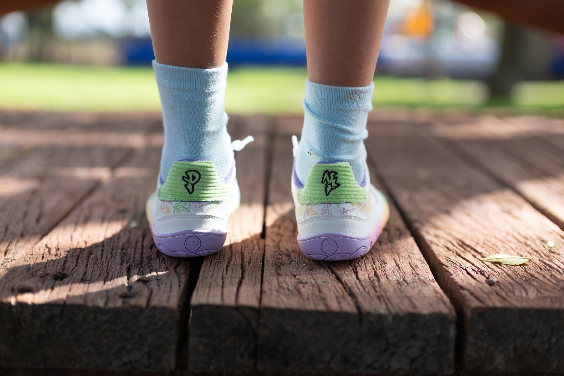 White barefoot sneakers from PaperKrane with a zero drop rainbow sole, and rainbow origami cranes printed on them. 
Seen from the back, there's green corduroy with PK logo details. This pair is being worn by a child with white socks on. On a wooden bridge.