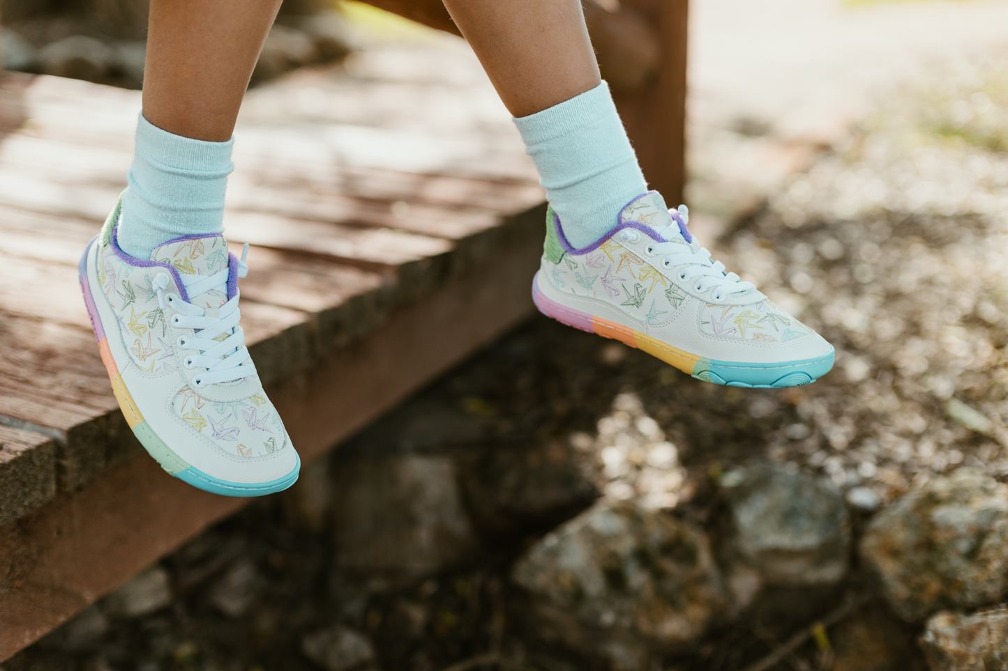 White barefoot sneakers from PaperKrane with a zero drop rainbow sole, and rainbow origami cranes printed on them. 
They have elastic laces and are worn by a child, with legs dangling off the side of a wooden structure.