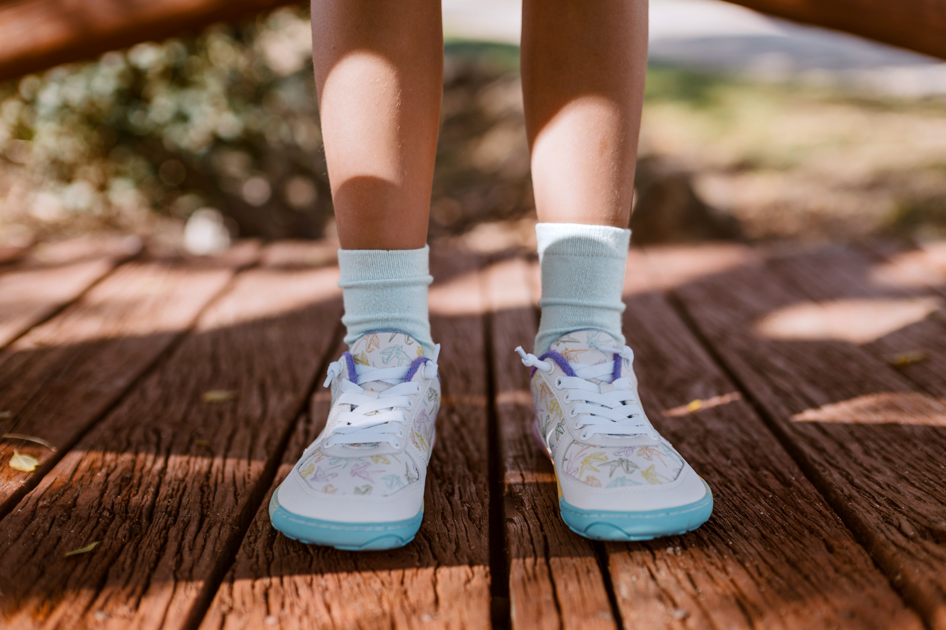 White barefoot sneakers from PaperKrane with a foot-shaped rainbow sole, and rainbow origami cranes printed on them. 
Seen from the front. This pair is being worn by a child with white socks on. On a wooden bridge.