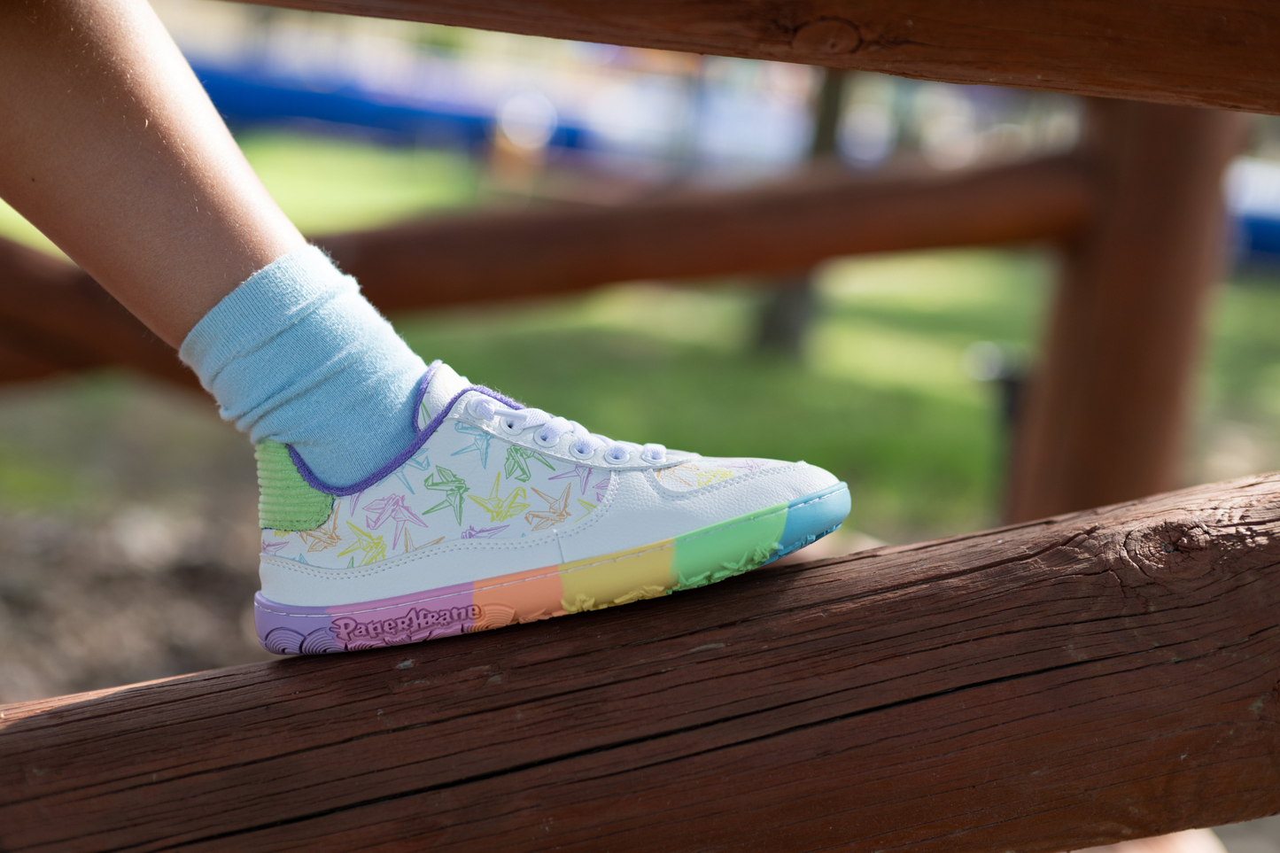 White barefoot sneakers from PaperKrane with a zero drop rainbow sole, and rainbow origami cranes printed on them. 
Seen from the side on a child's leg resting on a brown log.