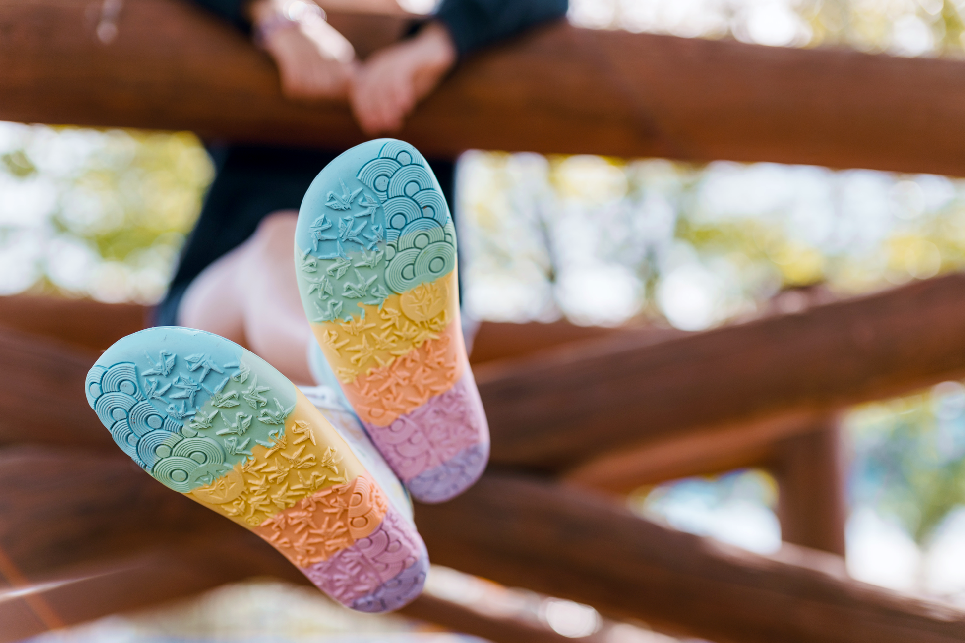 White barefoot sneakers from PaperKrane with foot-shaped rainbow soles. 
Seen from below, this pair is being worn by a child with white socks on. On a wooden structure.