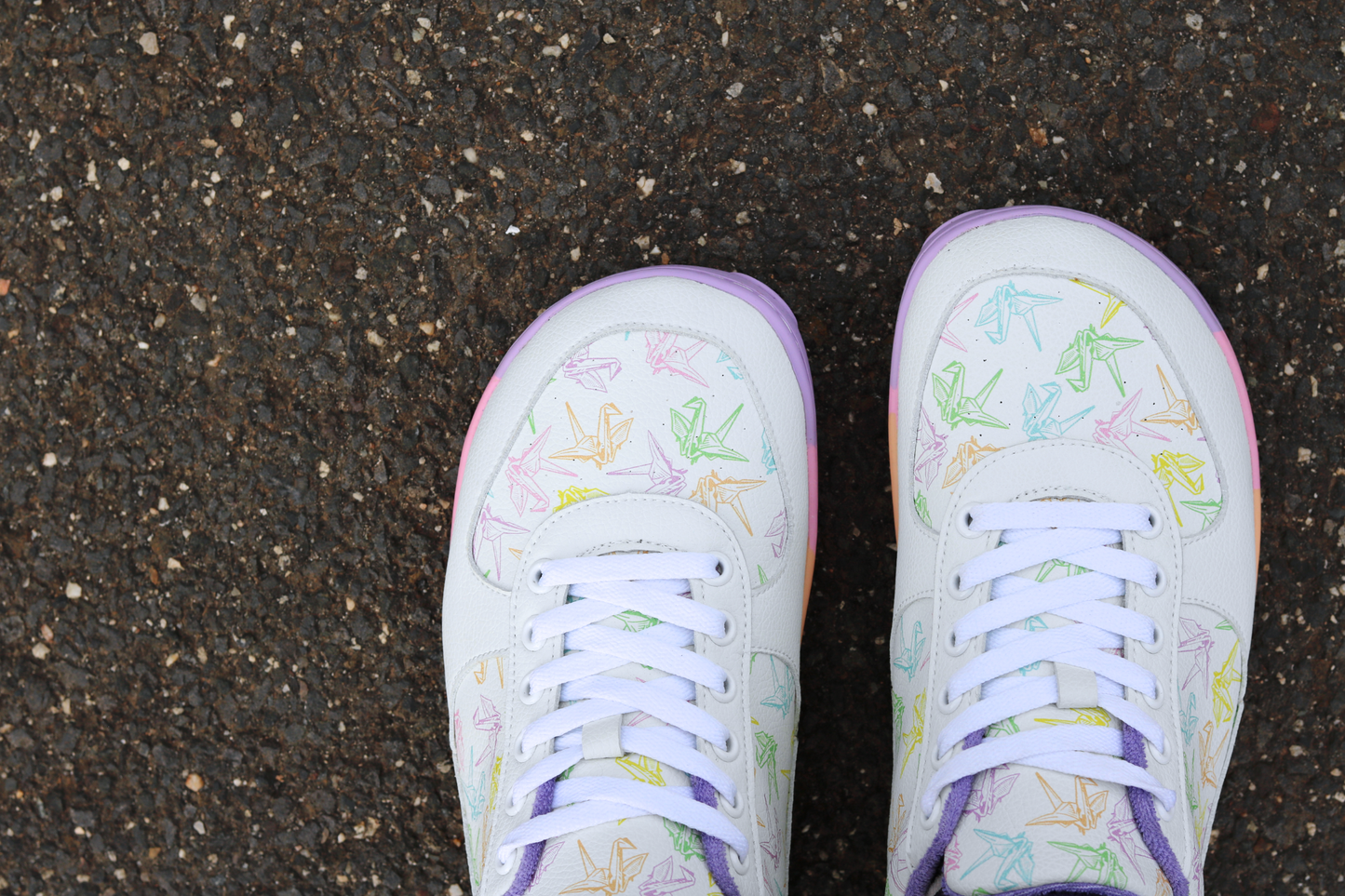 White barefoot sneakers from PaperKrane with a foot-shaped rainbow sole, and rainbow origami cranes printed on them. The lining is purple terry cotton. The view is from above the toe box, on asphalt. 