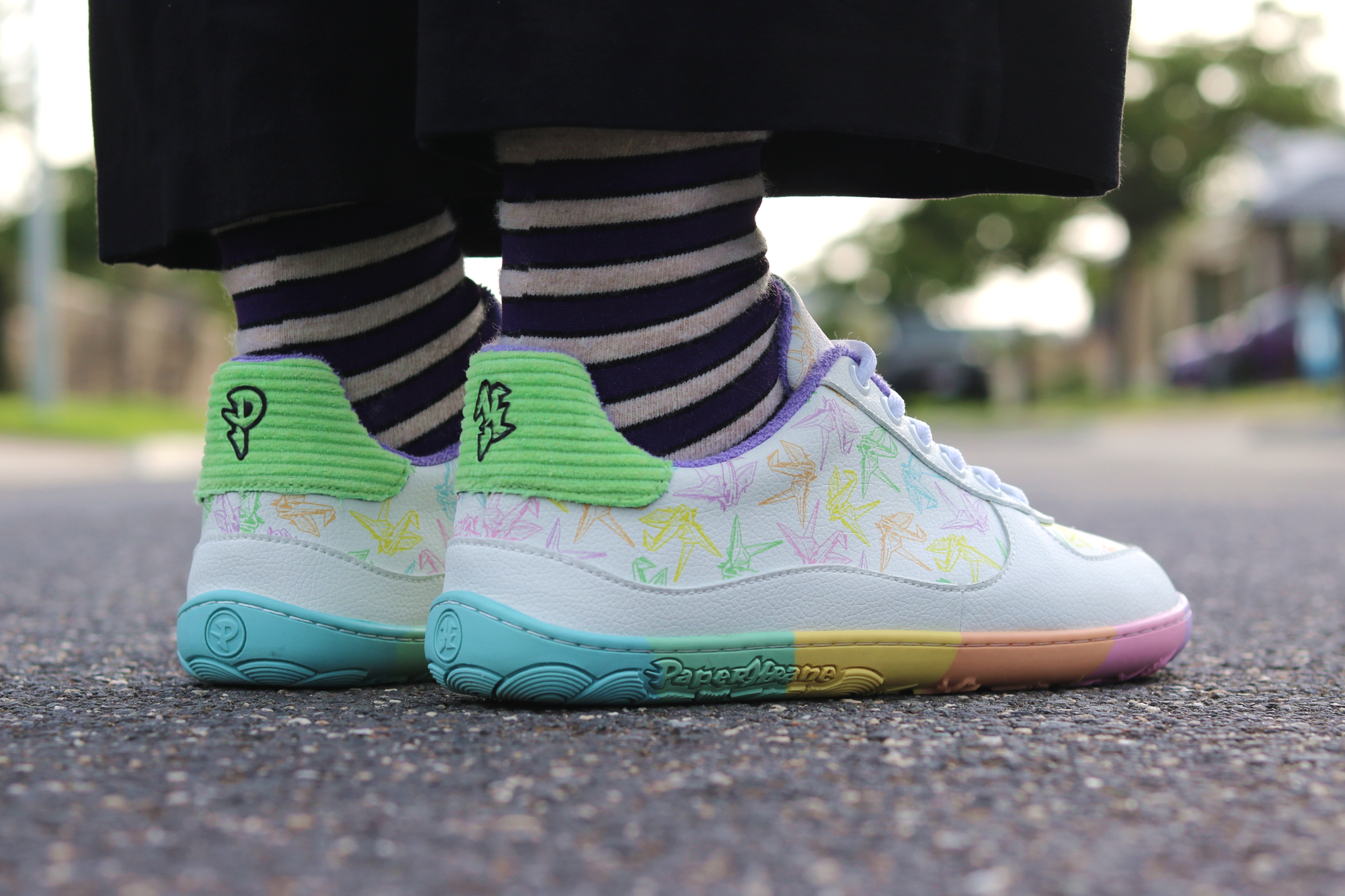 White barefoot sneakers from PaperKrane with a zero drop rainbow sole, and rainbow origami cranes printed on them. Worn by a person with purple striped merino socks, and black pants, on a road. 