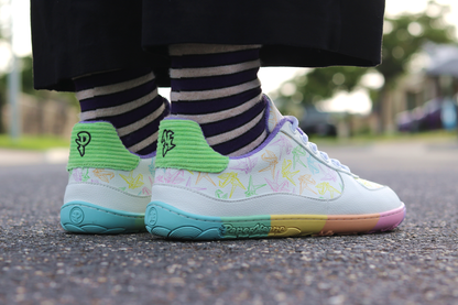 White barefoot sneakers from PaperKrane with a zero drop rainbow sole, and rainbow origami cranes printed on them. Worn by a person with purple striped merino socks, and black pants, on a road. 