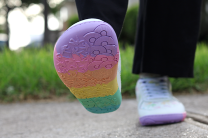 White barefoot sneakers from PaperKrane with a foot-shaped rainbow sole, worn by a person with black pants on a path in front of grass. One foot is lifted so you can see the sole and toe box shape. 