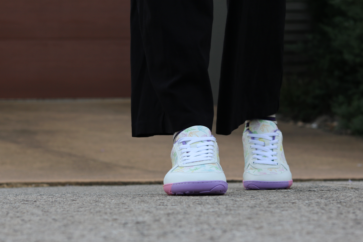 White barefoot sneakers from PaperKrane with a foot-shaped rainbow sole, and rainbow origami cranes printed on them. They're on a person with black pants on a footpath.