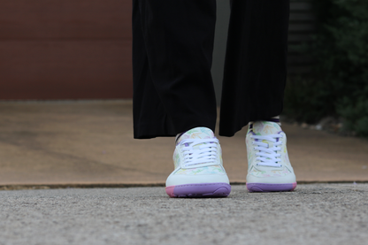 White barefoot sneakers from PaperKrane with a foot-shaped rainbow sole, and rainbow origami cranes printed on them. They're on a person with black pants on a footpath.