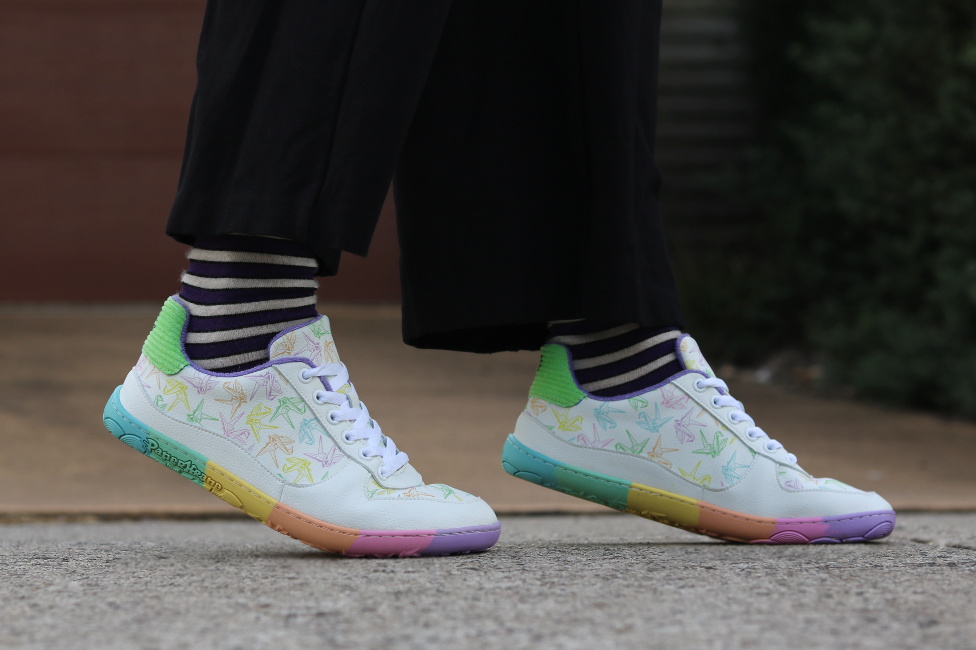 White barefoot sneakers from PaperKrane with a flexible foot-shaped rainbow sole, and rainbow origami cranes printed on them. 
Worn by a person with purple and beige striped merino socks from Roam Barefoot. Raised up on their toes. 