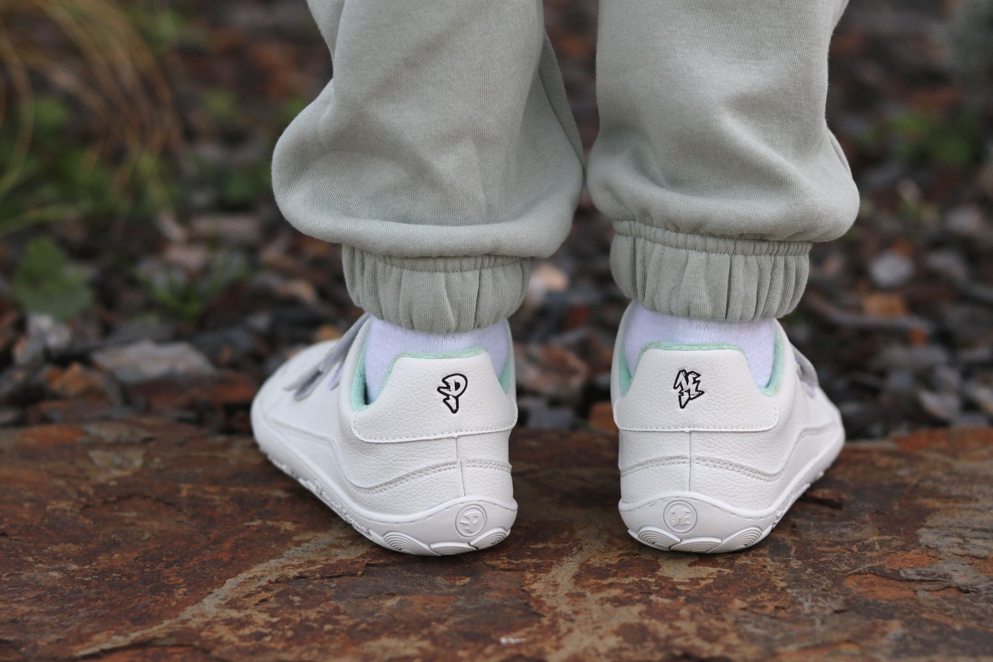White low-top sneakers with velcro straps, worn with light grey sweatpants, shown from the back on a person standing on a rustic stone surface outdoors. There is a black P and K logo on the back of the heels. 