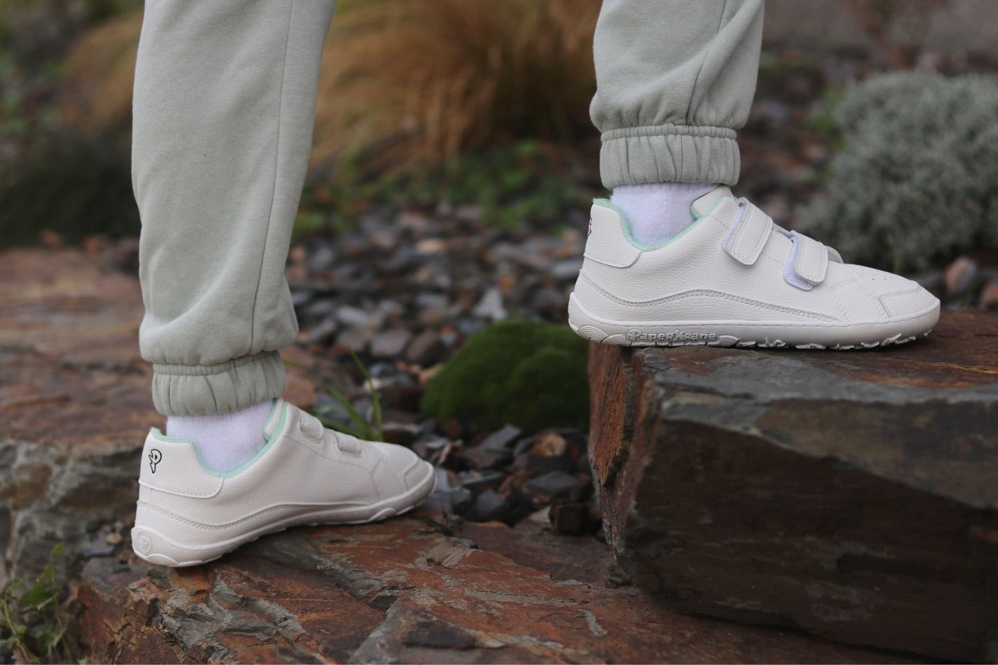 A child wearing light grey jogger pants and white vegan leather low-top velcro barefoot sneakers from PaperKrane with white socks, standing outdoors on stone steps with rocks and plants in the background.