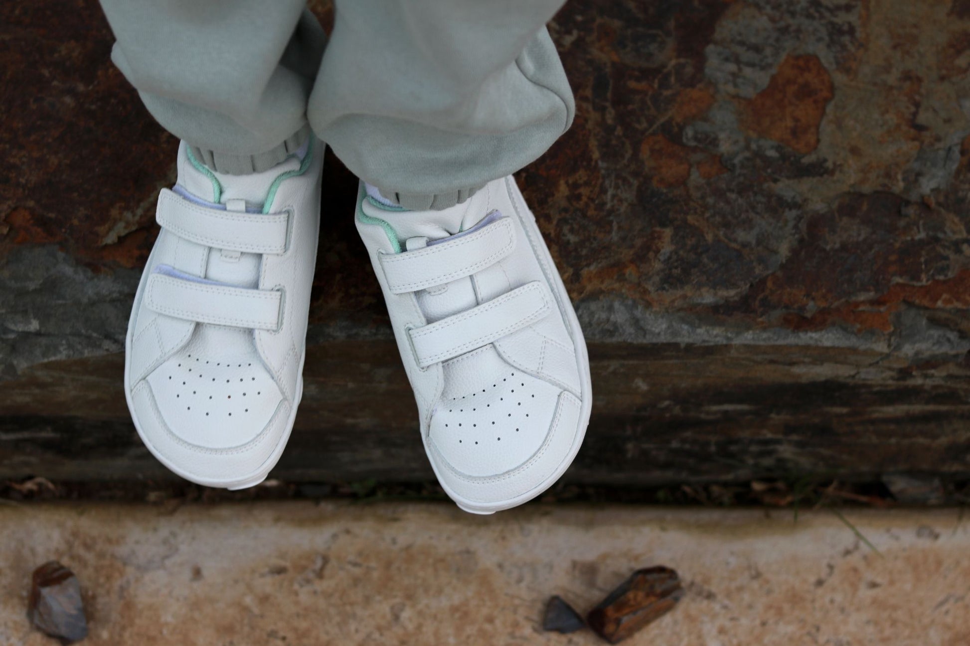 White vegan leather foot-shaped low-top sneakers with double hook-and-loop velcro straps, shown on a child's feet wearing light grey sweatpants, standing on a rustic stone surface.