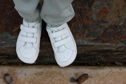 White vegan leather foot-shaped low-top sneakers with double hook-and-loop velcro straps, shown on a child's feet wearing light grey sweatpants, standing on a rustic stone surface.