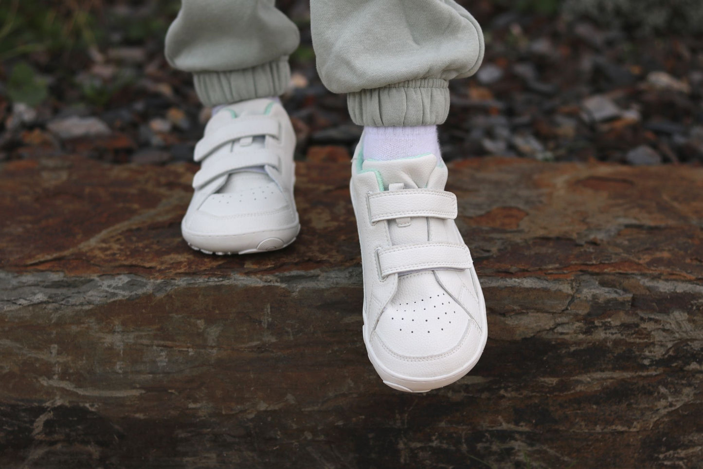 A close-up of a child's feet wearing white vegan low-top barefoot PaperKrane sneakers with velcro straps, standing on a large brown stone outdoors.