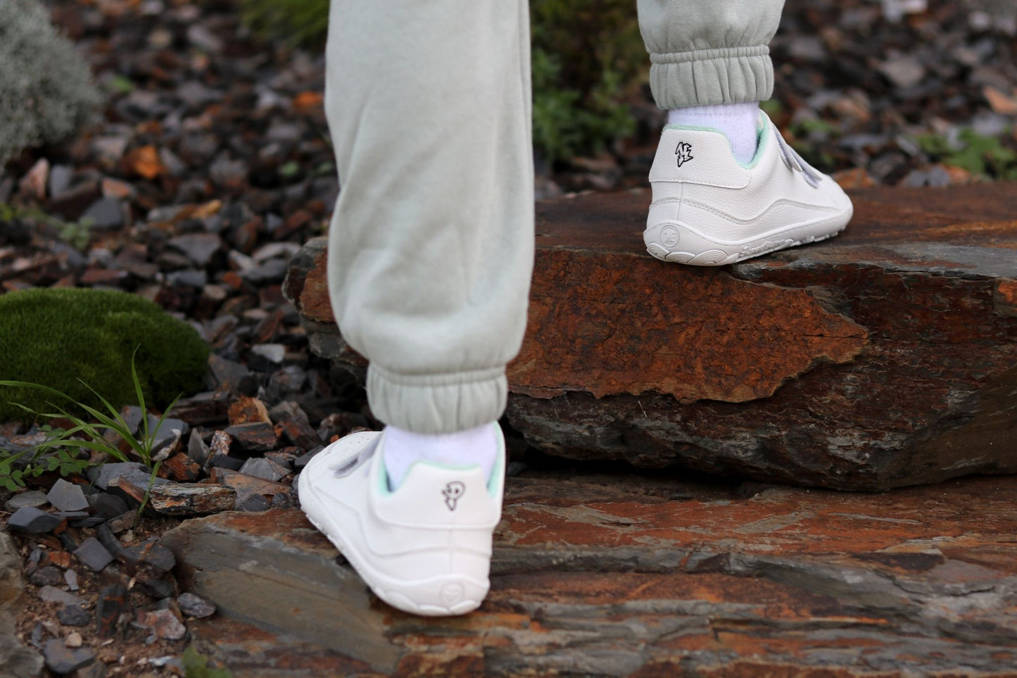 White vegan velcro sneakers from PaperKrane (PK) with white socks and light grey sweatpants, shown on a person stepping onto a large brown rock outdoors.