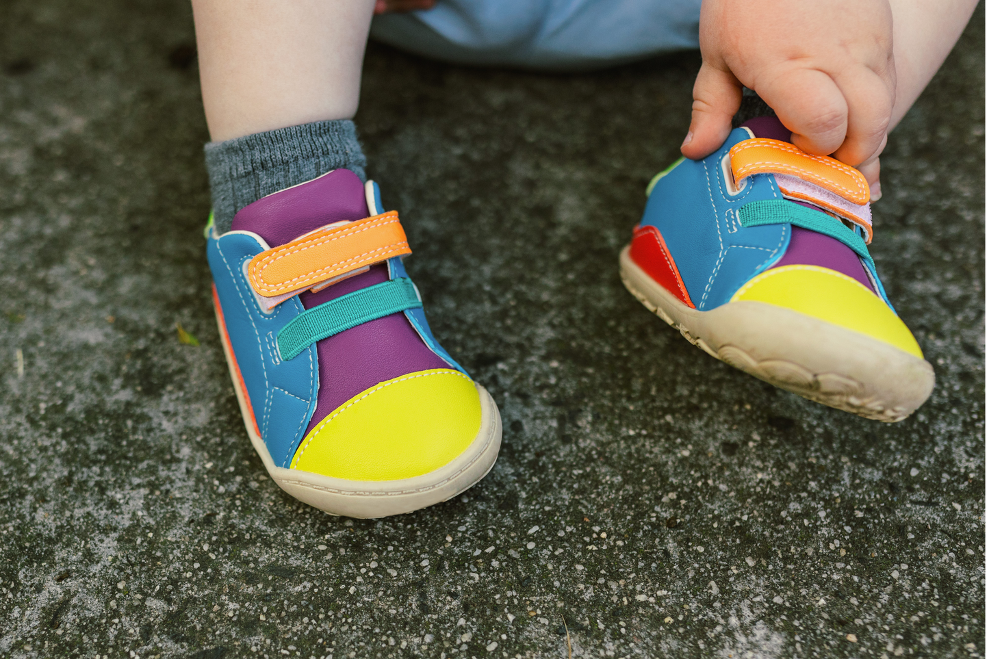 A TODDLER IN VEGGIE MIDI SNEAKERS, THEY HAVE BRIGHT YELLOW TOES, PURPLE TONGUE, ORANGE AND TEAL STRAPS, BLUE SIDES, AND BEIGE SOLES